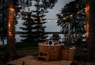 Glampingplek aan het meer met mensen in een houten hottub buiten en sfeervolle lichtjes tussen de bomen.