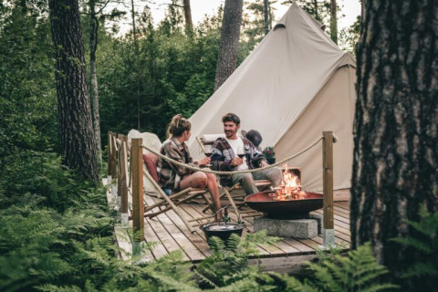 Twee mensen ontspannen bij een kampvuur voor een tent op een houten terras bij Nordic Woods Wildernis Glamping Zweden.