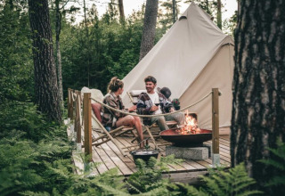 Twee mensen ontspannen bij een kampvuur voor een tent op een houten terras bij Nordic Woods Wildernis Glamping Zweden.