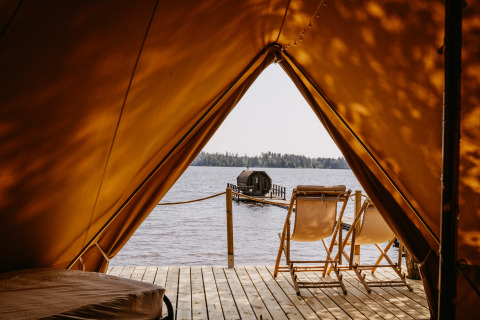Uitzicht vanuit tent op meer, twee stoelen op houten terras en sauna op steiger, Nordic Woods Wildernis Glamping Zweden
