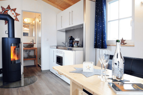 Modern glamping accommodation with wood ceiling, cozy fireplace, kitchenette, dining table, and bathroom.