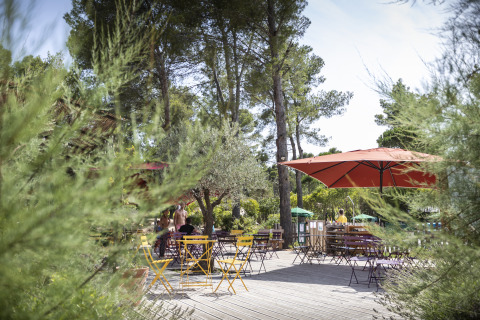 Terrazza all'aperto immersa nel verde con tavoli, sedie e ombrelloni a Huttopia Fontvieille – Glamping Provence.