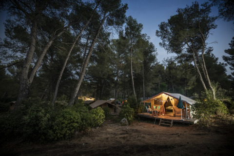 Huttopia Fontvieille – Glamping Provence: cozy illuminated tent in tranquil forest during dusk, outdoor camping.