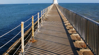 A pathway with railings leads to a lighthouse by the sea near L'Aiguillon sur Mer, France, under a blue sky.