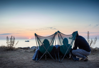 Three people relaxing in camping chairs at the beach while watching a sunset at Huttopia Noirmoutier - Glamping Vendée.