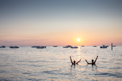 Sunset at the beach with two people swimming, boats in the distance at Huttopia Noirmoutier - Glamping Vendée.