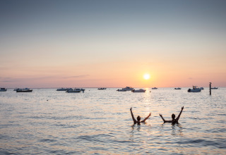 Sunset at the beach with two people swimming, boats in the distance at Huttopia Noirmoutier - Glamping Vendée.
