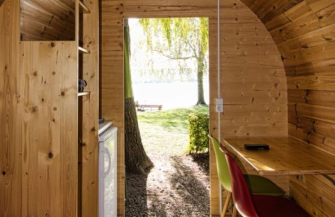 Interiør af et træpod med skrivebord og stole, åbent ud til grønne træer og sø ved Camping Markelfingen.