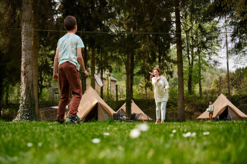 Familie spiller badminton foran tipitelte i grøn skov ved Camping de Marcourt, Belgisk Luxemburg.