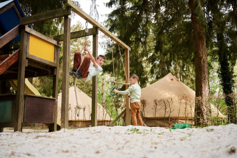 Twee kinderen spelen op een speeltuin met tipitenten op Camping de Marcourt in Belgisch Luxemburg op de achtergrond.