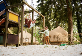 Twee kinderen spelen op een speeltuin met tipitenten op Camping de Marcourt in Belgisch Luxemburg op de achtergrond.