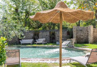 Tuinzwembad met waterval, ligstoelen en rieten parasol bij PROCIDA Camp & Resort - Luxe airstreams glamping.