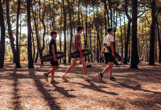 Three young people carrying longboards walk through a sunlit pine forest at Surf Tribe in Biscarrosse, France.