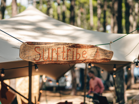 Wooden 'Surftribe' sign hanging at a glamping site in Biscarrosse, France, surrounded by a pine forest.