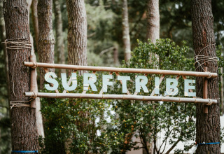 Holzschild mit der Aufschrift 'SURFTRIBE' zwischen Bäumen, Glamping-Unterkunft in Biscarrosse, Frankreich.