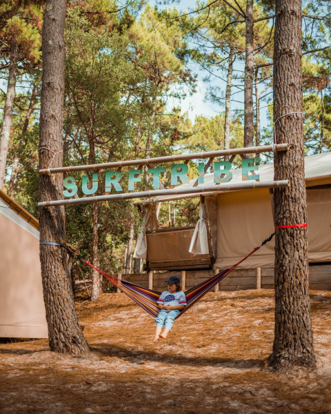 Kind in einer Hängematte unter dem Schild 'SURFTRIBE' bei Glamping-Lodges in Biscarrosse, Frankreich.