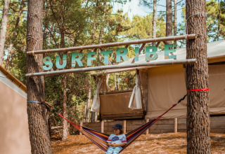 Barn i en hængekøje under skiltet 'SURFTRIBE' blandt træer ved glamping i Biscarrosse, Frankrig.