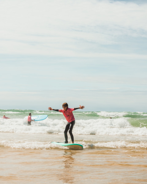 Barn surfer på lavt vand ved stranden i Biscarrosse med flere surfere i baggrunden, under solrig himmel.