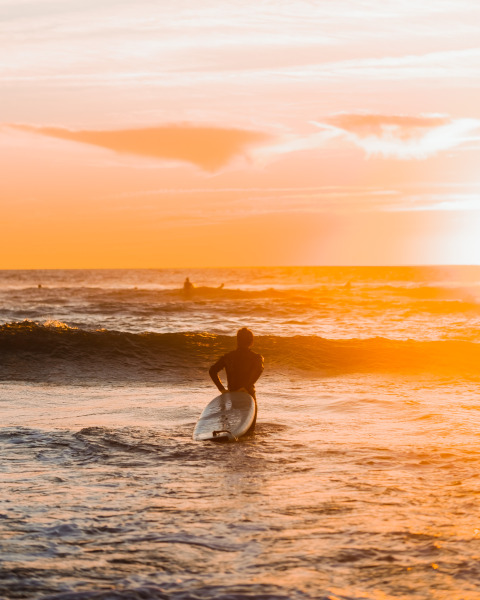 Surfeur entrant dans la mer au coucher du soleil près de Surf Tribe à Biscarrosse, expérience de glamping unique.