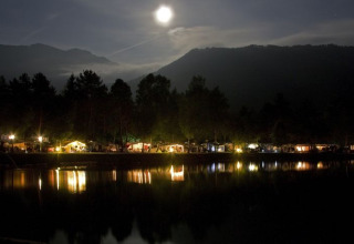 Vista nocturna de glamping en Camping Rosental Rož, alojamientos iluminados junto a un lago bajo la luna llena.
