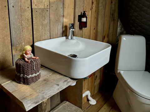 Baño rústico con paredes de madera, lavabo, inodoro y muñeca sobre papel higiénico en Brokkestøylen - Domes in Agder.