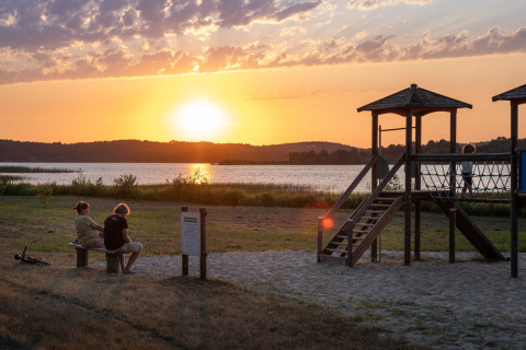 Sunset by the lake at Huttopia Lac d’Aureilhan – Mimizan glamping with playground and two people on benches.