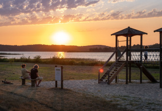 Puesta de sol junto al lago en Huttopia Lac d’Aureilhan – Mimizan glamping con área de juegos y bancos.