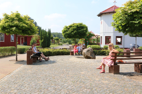 Menschen sitzen auf Bänken in Campingpark Buntspecht-Pods Brandenburg an einem sonnigen, begrünten Platz.
