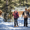 Two people snowshoeing on a trail near wooden cabins at Huttopia Vanoise – Champagny Winter glamping site.