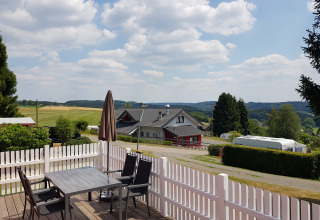 Vue depuis Camping im Bergischen Land avec terrasse, chaises, clôture blanche et caravane dans la verdure.