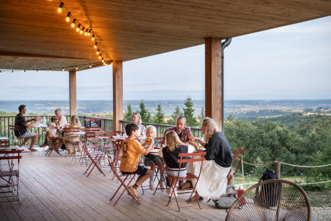 Familien und Freunde genießen ein Essen auf der Terrasse von Huttopia Pays de Condrieu mit Panoramablick auf die Natur.