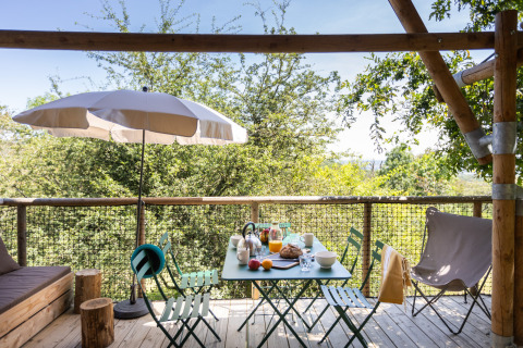 Udendørs spisebord på en træterrasse med paraply og skovudsigt på Village Huttopia Pays de Condrieu glamping.