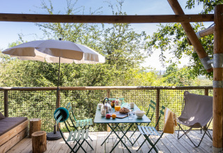 Udendørs spisebord på en træterrasse med paraply og skovudsigt på Village Huttopia Pays de Condrieu glamping.