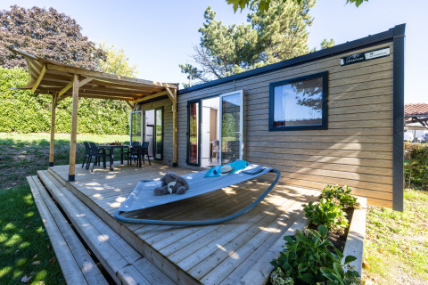 Holzterrasse mit Liegestuhl und Esstisch vor einem Glamping-Mobilheim bei Camping La Grappe Fleurie im Grünen.