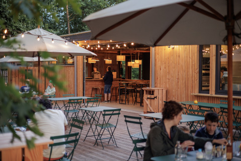 Zona de comedor exterior con mesas verdes en Camping Huttopia De Veluwe, luces cálidas al atardecer.