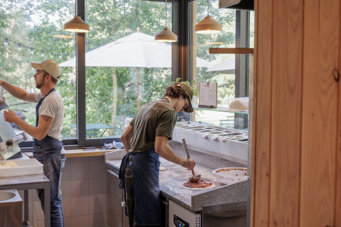 Two chefs prepare pizza in a bright kitchen with large windows at Camping Huttopia De Veluwe in Gelderland.