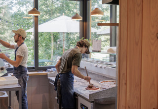 To kokke forbereder pizza i et lyst køkken med store vinduer på Camping Huttopia De Veluwe i Gelderland.
