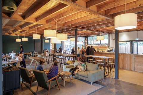 Indoor lounge area at Camping Huttopia De Veluwe, showing guests relaxing among wooden beams and lights.