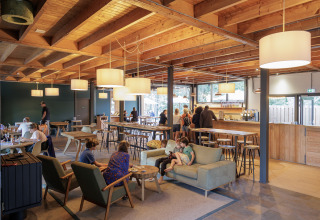 Indoor lounge area at Camping Huttopia De Veluwe, showing guests relaxing among wooden beams and lights.