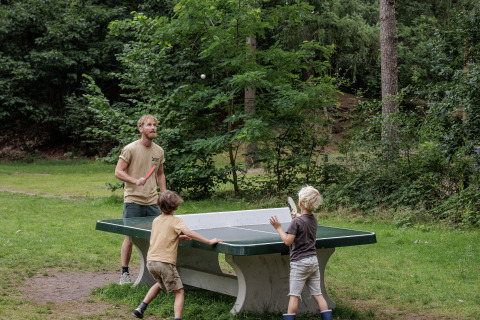 Een man en twee kinderen spelen buiten tafeltennis bij Camping Huttopia De Veluwe, omringd door bos.