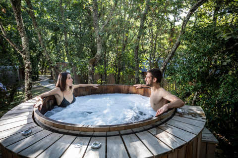 Two people relaxing in a wooden outdoor hot tub surrounded by trees at Village Huttopia Sud Ardèche – Glamping Ardèche.