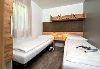 Alloggio glamping moderno con due letti singoli, mensola centrale e luce naturale dalla finestra.