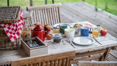Desayuno al aire libre con cesta de picnic, café, jugo, pan y fruta en mesa de madera en glamping.