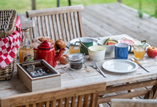 Desayuno al aire libre con cesta de picnic, café, jugo, pan y fruta en mesa de madera en glamping.