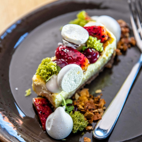 Stijlvol dessert met taartbodem, verse bessen, groene cakekruimels en witte meringue op donker bord.