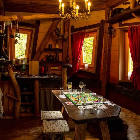 Gemütliche Holzhütte von Boomhut - Dierentuin Schwerin mit Brettspiel, rustikalen Möbeln und roten Vorhängen.