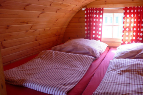 Interno di alloggio glamping in legno con due letti singoli, lenzuola a righe e tende rosse a pois bianchi.