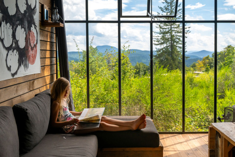 Ein Mädchen liest ein Buch auf einem Sofa in einer Nutchel Cabin mit Panoramablick auf die Natur.
