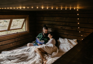 Couple relaxing in bed reading a book inside Nutchel Cabins - Tiny Houses Elzas glamping cabin.