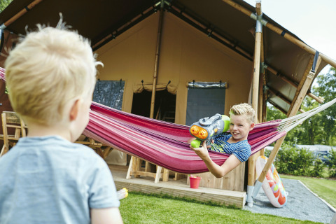 Junge zielt mit Wasserpistole auf anderen Jungen in Hängematte vor Glamping-Zelt bei Recreatiepark TerSpegelt.